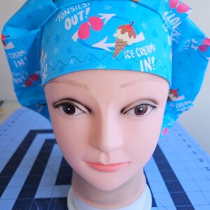 May include: A light blue surgical scrub cap with a repeating pattern of ice cream cones, tonsils, and stars. The cap has the phrases "Tonsils Out!" and "Ice Cream In!" printed on it. The cap is designed to be worn in a medical setting.