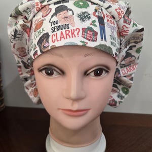 May include: A white scrub cap featuring a repeating pattern of Christmas-themed illustrations and text including the phrase YOU SERIOUS CLARK?, a turkey, a dog, a gift box labeled SHE WRAPPED UP HER DAMN CAT, and various holiday icons.