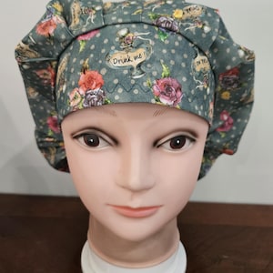 May include: A scrub cap in a muted green fabric featuring a pattern of polka dots, colorful roses, and illustrations of glass bottles with the text Drink me! printed on them. The cap has a pleated front band and a gathered back for a secure fit.