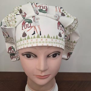 May include: A white scrub cap featuring a repeating pattern of a person in a bathrobe holding a hose, a pile of brown waste, and the text "Shitter's FULL" in red and green. The cap includes a green pine tree border and a pleated front band.