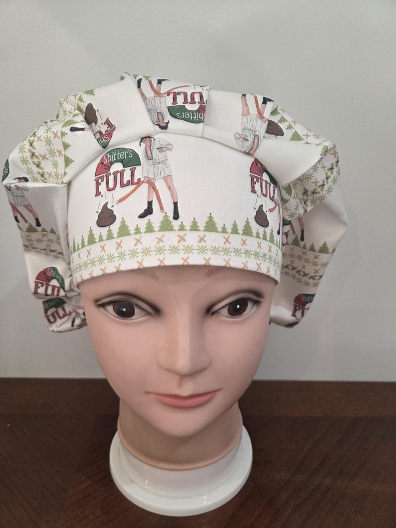 May include: A white scrub cap featuring a repeating pattern of a person in a bathrobe holding a hose, a pile of brown waste, and the text "Shitter's FULL" in red and green. The cap includes a green pine tree border and a pleated front band.