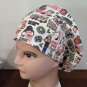 May include: A white scrub cap featuring a colorful all-over print of characters, vehicles, and holiday-themed objects from a Christmas movie. The fabric includes text such as CLARK?, FULL, YOU SERIOUS, DON'T HOG, and SAVE THE NECK.