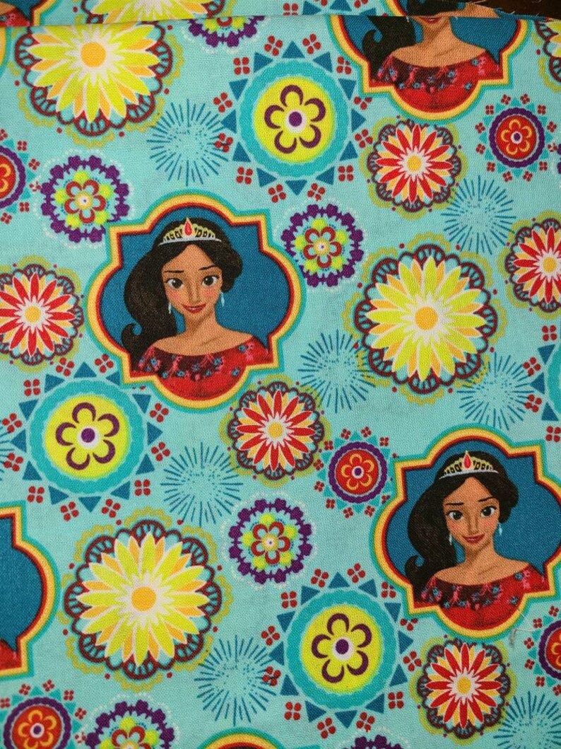 May include: A turquoise fabric with a repeating pattern of colorful flowers, stars, and a princess with a crown. The princess is wearing a red dress and has dark hair.