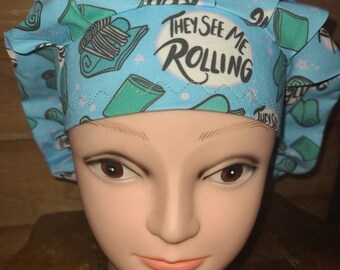 May include: A blue scrub hat with a white background and a repeating pattern of teal and white rolled up towels. The hat has the text "They See Me Rolling" in white.