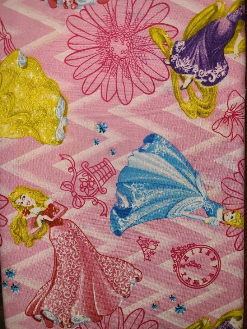 May include: Pink fabric with a repeating pattern of Disney princesses. The princesses are Cinderella, Rapunzel, and Aurora. The fabric also features a clock, a flower, and a birdcage.