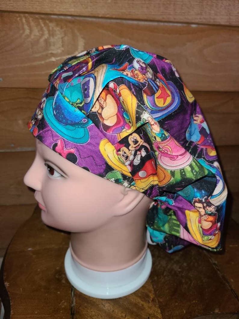 May include: A purple, green, and black scrub hat with a colorful cartoon print featuring Mickey Mouse, Minnie Mouse, and other Disney characters.