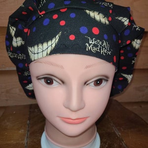 May include: Black scrub hat with red and blue polka dots and a white grinning mouth design. The hat has the text "We're All Mad Here" in white.