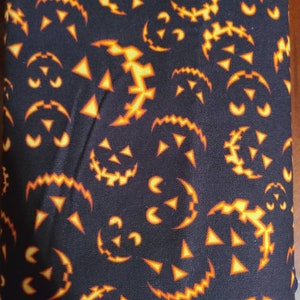 May include: Black fabric with a repeating pattern of orange jack-o'-lantern faces. The faces are outlined in black and have a jagged, toothy grin.