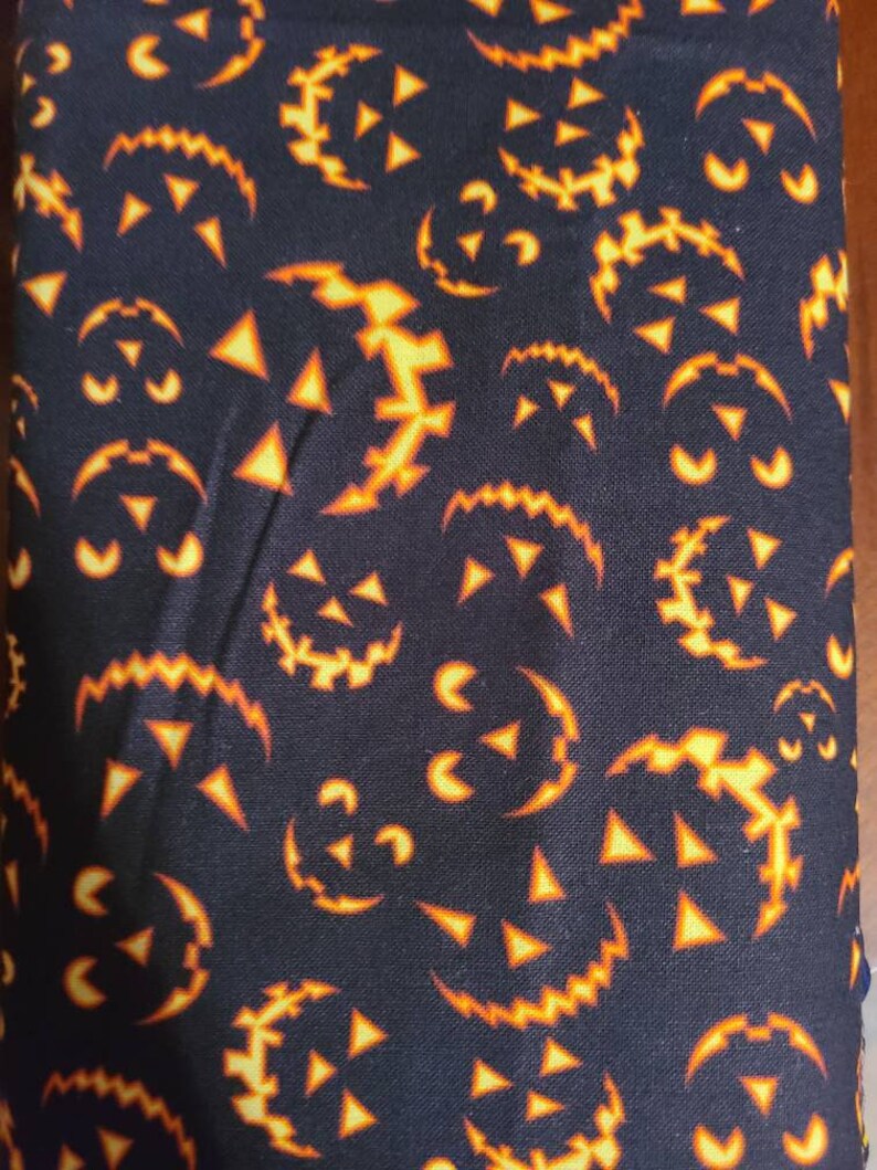 May include: Black fabric with a repeating pattern of orange jack-o'-lantern faces. The faces are outlined in black and have a jagged, toothy grin.