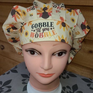 May include: A beige scrub hat with a Thanksgiving theme. The hat has a pattern of turkeys, sunflowers, and fall leaves. The text "Gobble 'Til You Wobble" is embroidered on the hat.