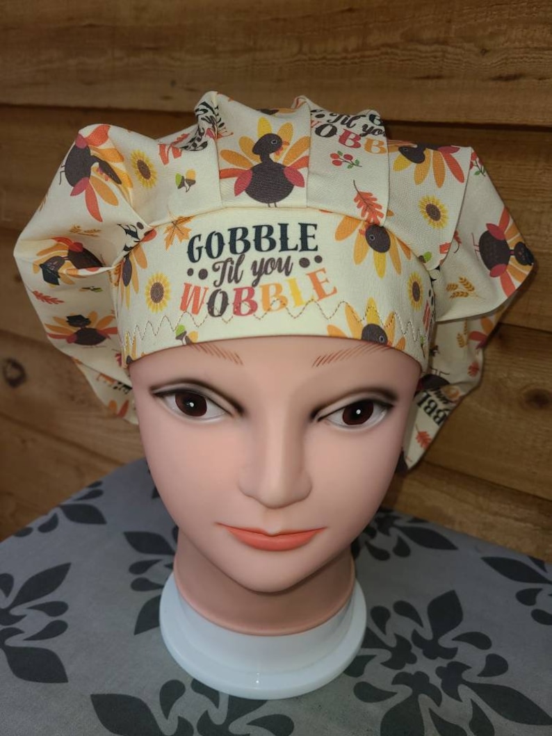 May include: A beige scrub hat with a Thanksgiving theme. The hat has a pattern of turkeys, sunflowers, and fall leaves. The text "Gobble 'Til You Wobble" is embroidered on the hat.