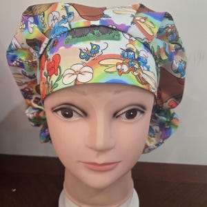 May include: A scrub cap featuring a colorful cartoon print of blue characters, butterflies, flowers, and rainbows against a light background. The fabric has a gathered design with a wide, structured front band and a pleated crown construction.