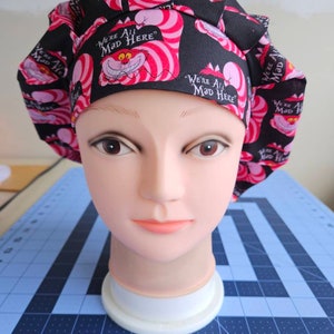 May include: Black scrub hat with a pink and white pattern featuring the Cheshire Cat from Alice in Wonderland. The hat has the text "We're All Mad Here" repeated on it.