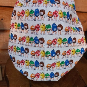 Pu&ograve; includere: Un berretto da lavoro bianco con un motivo ripetuto di personaggi M&M's colorati. I personaggi indossano tutti scarpe bianche e si tengono per mano. Lo sfondo &egrave; bianco.