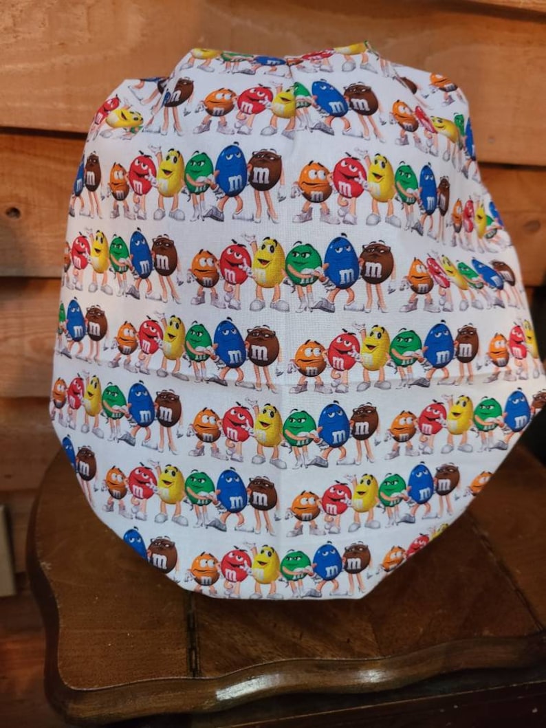Pu&ograve; includere: Un berretto da lavoro bianco con un motivo ripetuto di personaggi M&M's colorati. I personaggi indossano tutti scarpe bianche e si tengono per mano. Lo sfondo &egrave; bianco.
