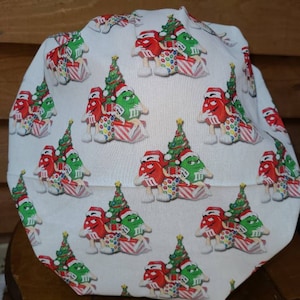 Pu&ograve; includere: Un berretto da lavoro bianco in tessuto con un motivo ripetuto di personaggi M&M's rossi e verdi che indossano cappelli da Babbo Natale e tengono regali di Natale. I personaggi sono in piedi davanti ad alberi di Natale verdi.