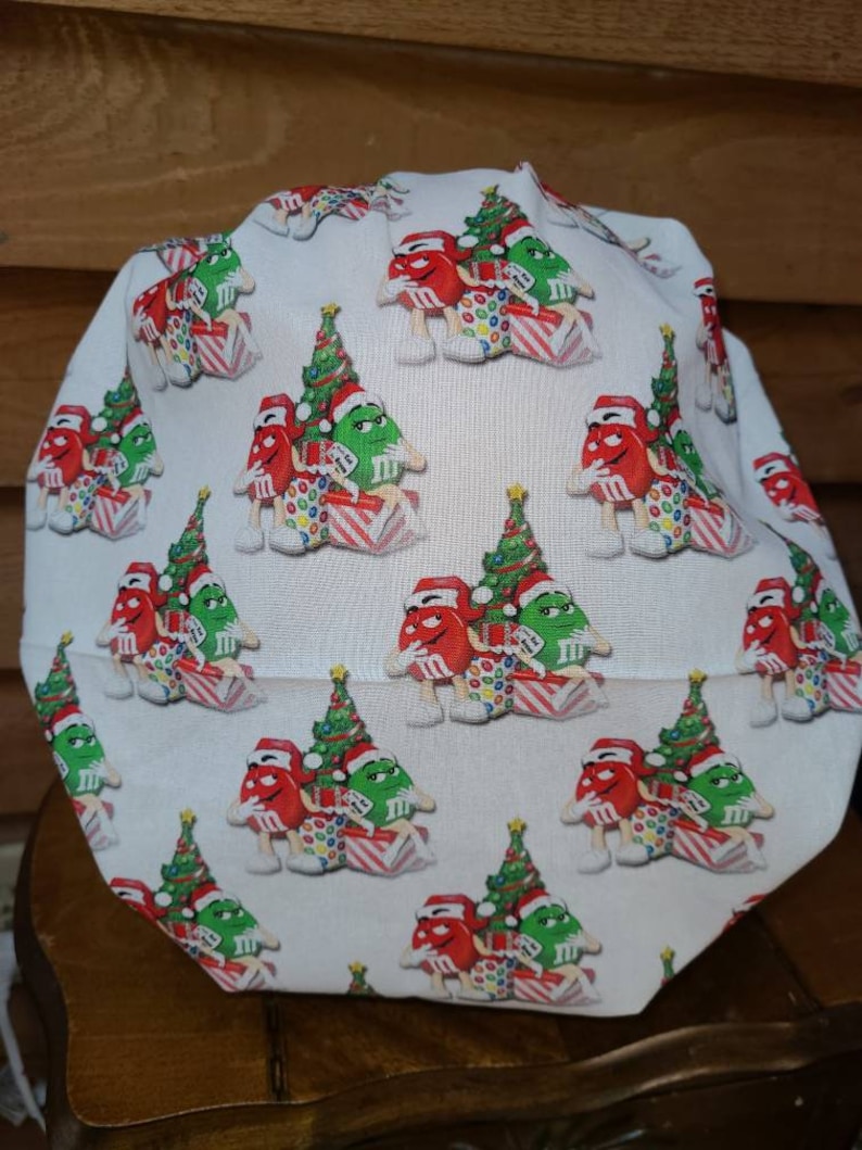 Pu&ograve; includere: Un berretto da lavoro bianco in tessuto con un motivo ripetuto di personaggi M&M's rossi e verdi che indossano cappelli da Babbo Natale e tengono regali di Natale. I personaggi sono in piedi davanti ad alberi di Natale verdi.