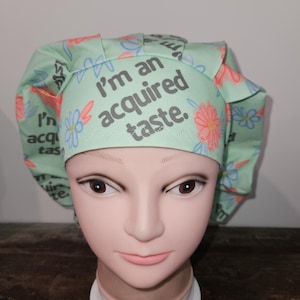 May include: A green scrub hat with a floral pattern and the text "I'm an acquired taste."