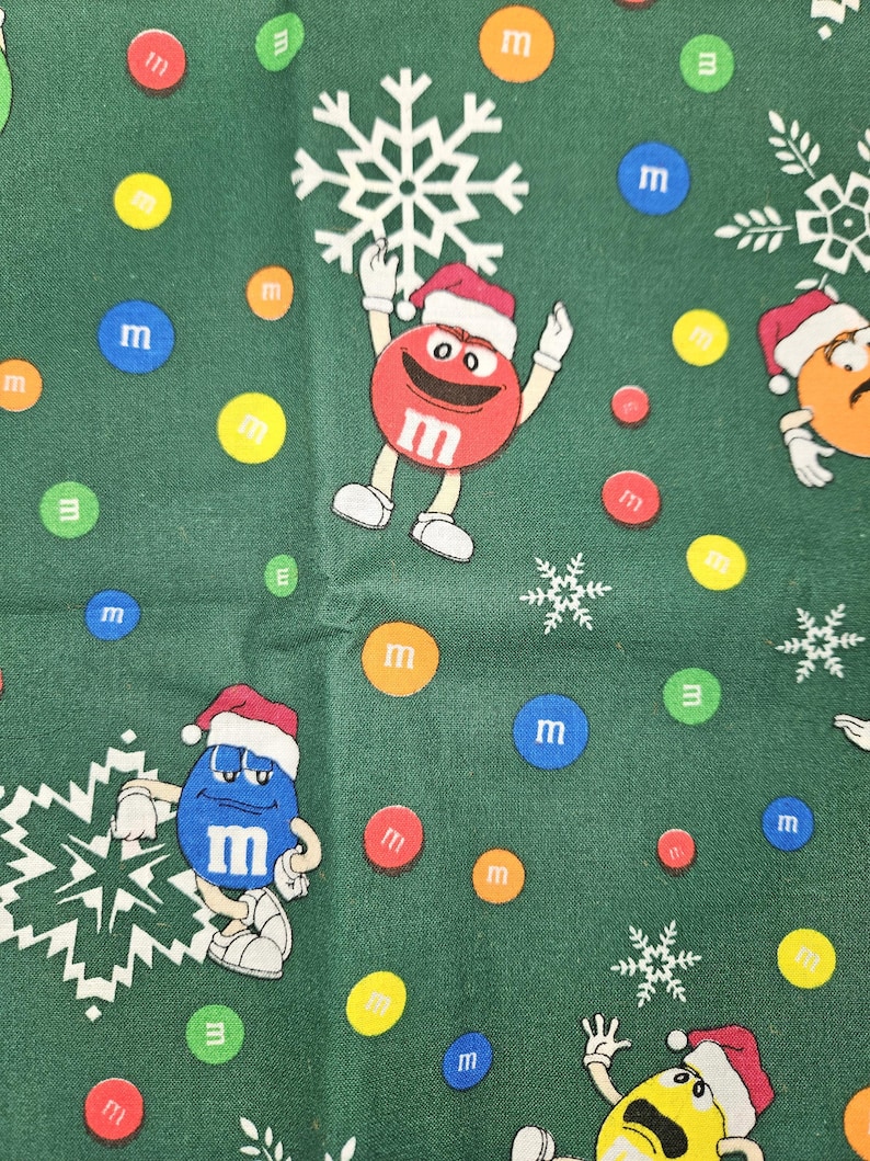 Pu&ograve; includere: Tessuto verde con un motivo ripetuto di M&M's che indossano cappelli da Babbo Natale e tengono fiocchi di neve. Gli M&M's sono rossi, blu, gialli e arancioni. Ci sono anche M&M's sparsi in vari colori.