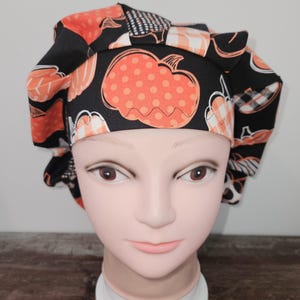 May include: A black scrub hat with a pumpkin print in orange and white. The hat has a gathered top and a tie at the back.