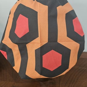 May include: A fabric item with a repeating geometric pattern of black, orange, and red hexagons. The item is folded, creating a rounded shape. The design is reminiscent of a classic carpet pattern.