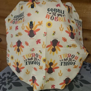 May include: A white fabric with a Thanksgiving theme. It features a repeating pattern of turkeys, sunflowers, wheat, and leaves. The text "Gobble Til You Wobble" is repeated throughout the fabric.