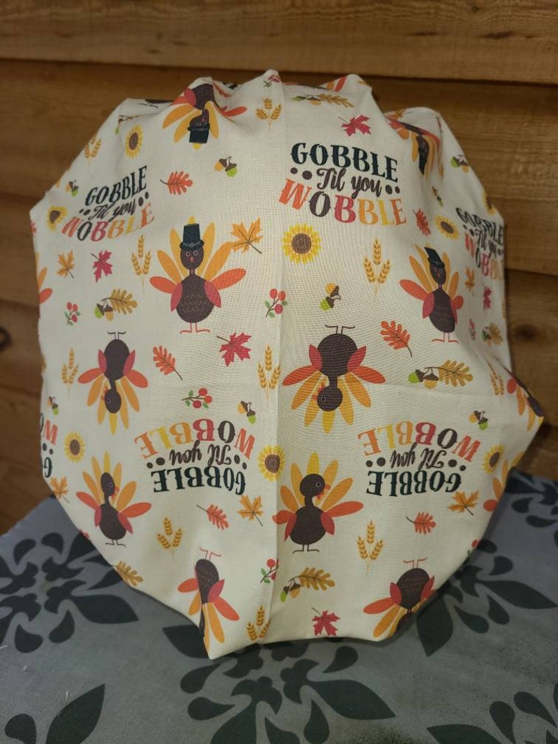 May include: A white fabric with a Thanksgiving theme. It features a repeating pattern of turkeys, sunflowers, wheat, and leaves. The text "Gobble Til You Wobble" is repeated throughout the fabric.