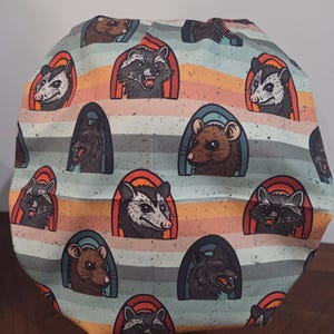 May include: A scrub cap featuring a repeating pattern of raccoons, opossums, rats, and wolves. Each animal is framed within a colorful arch against a background of horizontal stripes in shades of light blue, orange, and tan with a speckled texture.