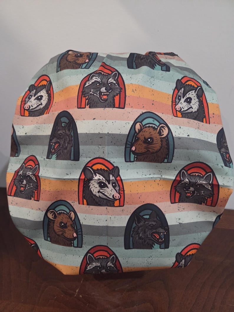 May include: A scrub cap featuring a repeating pattern of raccoons, opossums, rats, and wolves. Each animal is framed within a colorful arch against a background of horizontal stripes in shades of light blue, orange, and tan with a speckled texture.