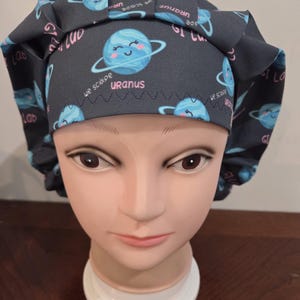 May include: A dark gray bouffant scrub cap featuring a repeating pattern of smiling blue planets with rings and the text "Uranus" and "we scope" in pink. The cap is designed to cover the hair.