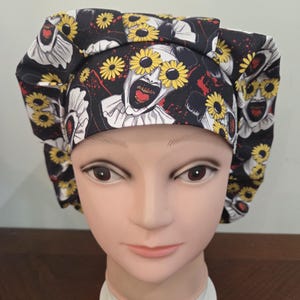 May include: A black scrub cap featuring a repeating pattern of a clown with a wide, open mouth, red lips, and yellow sunflower-shaped glasses. The fabric includes scattered red paint splatter accents against a dark background.
