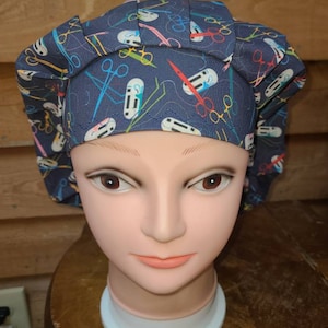 May include: A blue surgical scrub hat with a colorful pattern of scissors and medical tools. The hat is pleated and has a rounded shape.