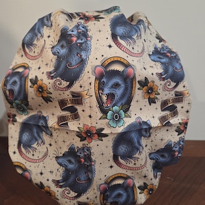 May include: A scrub cap featuring a cream-colored fabric with a repeating pattern of blue opossums, some carrying smaller opossums on their backs, interspersed with yellow and blue flowers, stars, and banners with the text AWESOME OPOSSUM.