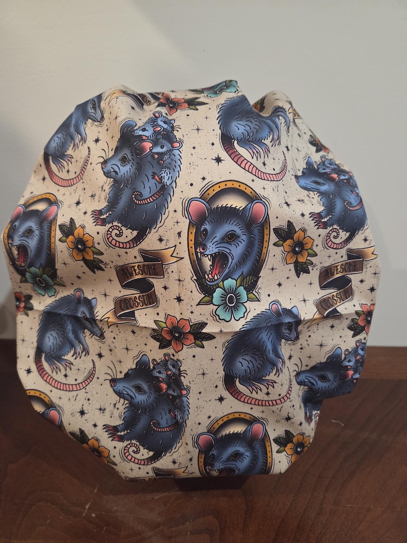 May include: A scrub cap featuring a cream-colored fabric with a repeating pattern of blue opossums, some carrying smaller opossums on their backs, interspersed with yellow and blue flowers, stars, and banners with the text AWESOME OPOSSUM.