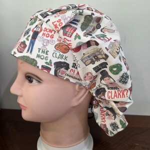 May include: A white scrub cap featuring a colorful Christmas-themed print with various illustrations and text including "Don't Hog The Clark Nog," "Serious Clark?," and "Aunt Bethany." The fabric has a pleated design and a small ChristieStitches label.