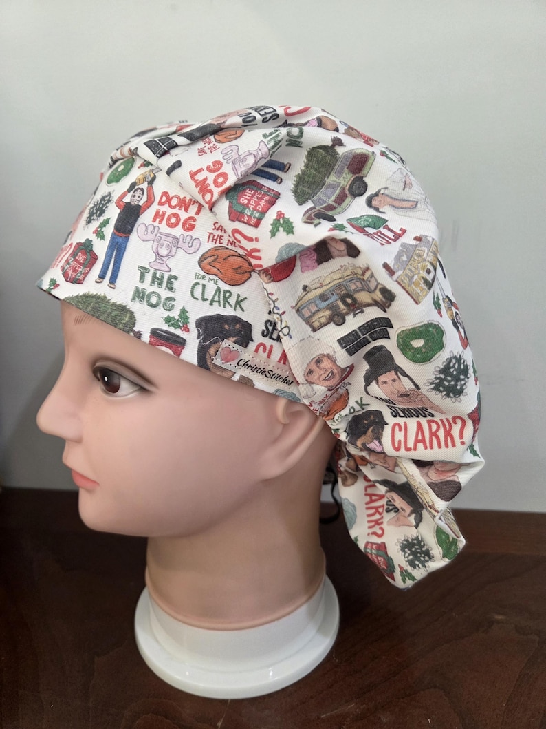 May include: A white scrub cap featuring a colorful Christmas-themed print with various illustrations and text including "Don't Hog The Clark Nog," "Serious Clark?," and "Aunt Bethany." The fabric has a pleated design and a small ChristieStitches label.