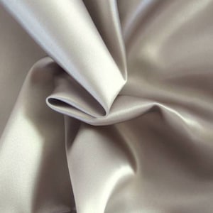May include: Close-up of a smooth, silky fabric in a light gray color. The fabric is draped and folded, creating soft curves and folds.