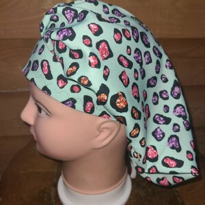 May include: A turquoise scrub hat with a pink, purple, and orange leopard print. The hat is made of a soft, comfortable fabric and has a tie at the back.