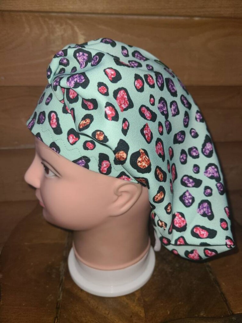 May include: A turquoise scrub hat with a pink, purple, and orange leopard print. The hat is made of a soft, comfortable fabric and has a tie at the back.