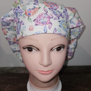May include: A white scrub cap with a colorful watercolor print of sea turtles. The turtles are in shades of pink, purple, blue, and green.