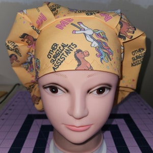 K&ouml;nnte beinhalten: Gelbe chirurgische M&uuml;tze mit einem Einhorn- und Pferde-Design. Die M&uuml;tze tr&auml;gt den Text "Other Surgical Assistants".