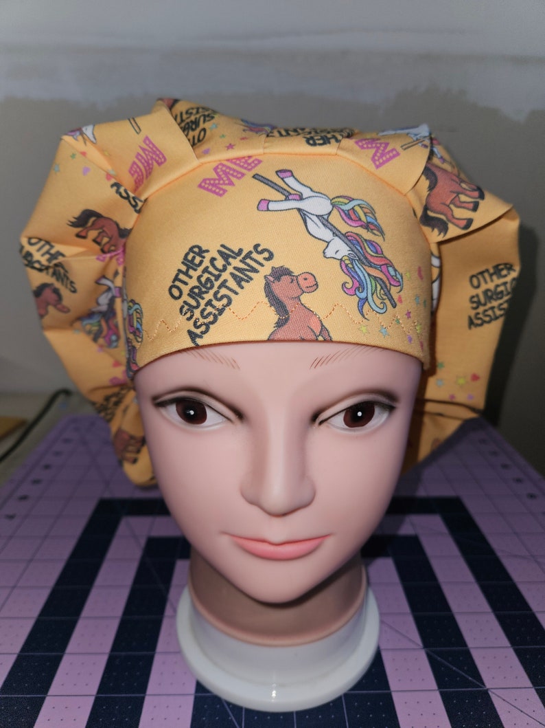 K&ouml;nnte beinhalten: Gelbe chirurgische M&uuml;tze mit einem Einhorn- und Pferde-Design. Die M&uuml;tze tr&auml;gt den Text "Other Surgical Assistants".