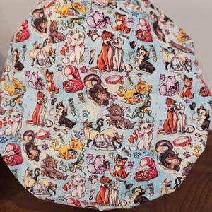 May include: A blue fabric scrub hat with a repeating pattern of cartoon cats from the Disney movie The Aristocats. The cats are in various poses and colors, including orange, white, black, and gray. The background is a light blue with white paw prints and hearts.