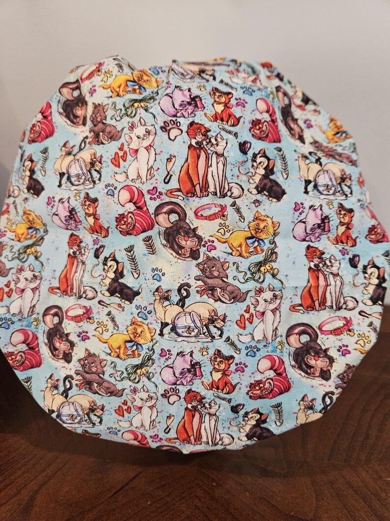 May include: A blue fabric scrub hat with a repeating pattern of cartoon cats from the Disney movie The Aristocats. The cats are in various poses and colors, including orange, white, black, and gray. The background is a light blue with white paw prints and hearts.
