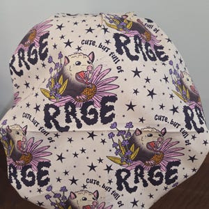 May include: A scrub cap with a cream background featuring a repeating pattern of opossums, purple flowers, and black stars. The text "cute, but full of RAGE" is printed in black lettering throughout the fabric design.