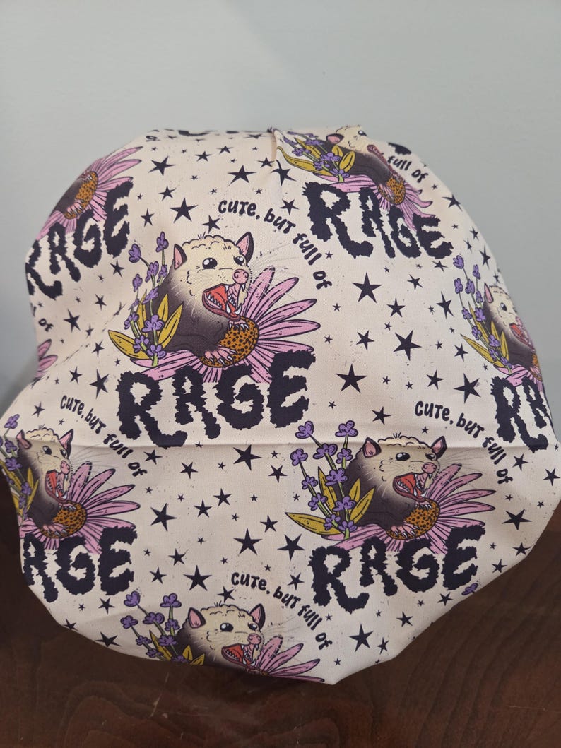 May include: A scrub cap with a cream background featuring a repeating pattern of opossums, purple flowers, and black stars. The text "cute, but full of RAGE" is printed in black lettering throughout the fabric design.