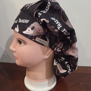 May include: A black scrub cap featuring a repeating pattern of gray opossums and the text A LITTLE TRASHY printed in white. The fabric cap has a gathered back design and a small white rectangular tag with the brand name ChristieStitches.