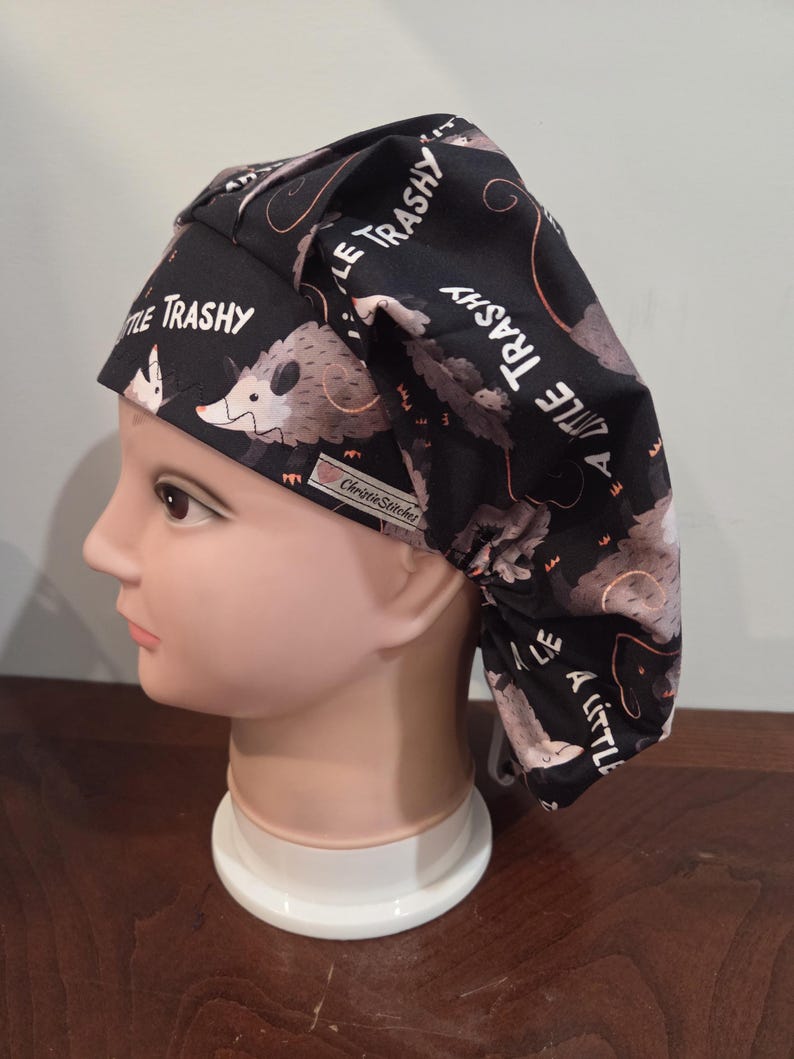 May include: A black scrub cap featuring a repeating pattern of gray opossums and the text A LITTLE TRASHY printed in white. The fabric cap has a gathered back design and a small white rectangular tag with the brand name ChristieStitches.
