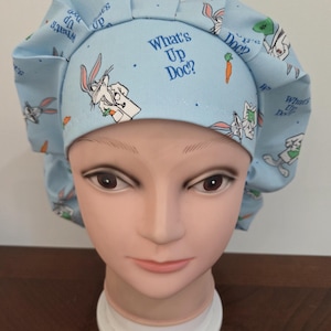 May include: A light blue scrub cap featuring Bugs Bunny illustrations and the phrase "What's Up Doc?". The cap has a gathered design and is decorated with carrots and Bugs Bunny in various poses.