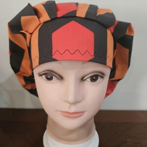 May include: A patterned scrub cap in orange, black, and red, displayed on a mannequin head. The cap features a geometric design with a red hexagon at the center and a black zigzag stitch along the brim. The fabric has an orange and black geometric pattern.
